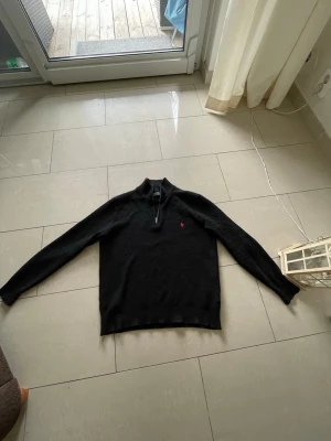 Svart stickad tröja från Polo Ralph Lauren - Svart stickad tröja från Polo Ralph Lauren med halv dragkedja och röd broderad logga på bröstet. Tröjan har ribbstickad krage, ärmslut och nederkant. Perfekt för lager på lager och klassisk stil.