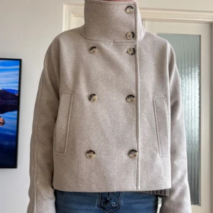 Beige kort ullrock med dubbelknäppning - Snygg beige kort rock med dubbelknäppning och hög krage. Rocken har stora knappar i marmorerad stil och två diskreta sidofickor. Tillverkad i ett tjockare material som ger en stilren och modern look. Perfekt för dig som vill ha en trendig och tidlös jacka.