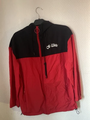 Röd och svart vindjacka Off-White - Storleks taggen har åkt av. Snygg vindjacka från Off-White i rött och svart med huva och dragkedja framtill. Jackan har vit Off-White-logga på bröstet och texten 'Impressionism' på ryggen. Två sidofickor med dragkedja och justerbara snören vid fållen. Perfekt för streetwear-stilen.