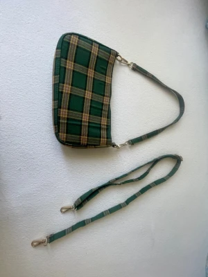 Grön rutig väska  - Snygg och trendig axelväska i grönt rutigt mönster perfekt till både vardag och sommarens outfits 🌿  Väskan har en justerbar axelrem samt en extra avtagbar rem, så du kan bära den på flera olika sätt (crossbody eller axelväska). Den har flera fack med dragkedja vilket gör den både praktisk och rymlig trots sin smidiga storlek. Insidan är fräsch och väskan är i fint skick. Perfekt för mobil, plånbok, nycklar och småsaker. Skickar snabbt!   