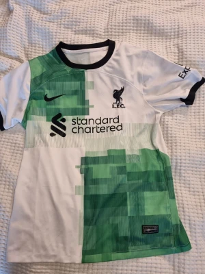 Liverpool FC bortatröja Nike grön/vit - Snygg Liverpool FC tröja från Nike i vitt med gröna grafiska mönster. Tröjan har svart rund halsringning, klubbmärke och Nike-logga på bröstet samt sponsortryck framtill. Fast pris, inga byten.