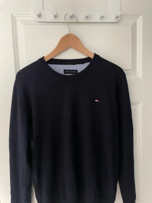 Mörkblå tröja från Tommy Hilfiger - Klassisk mörkblå tröja från Tommy Hilfiger med rund halsringning och diskret logga broderad på bröstet. Tröjan har ribbade muddar vid ärmslut och nederkant. Perfekt till jeans eller chinos för en clean look. Storlek M