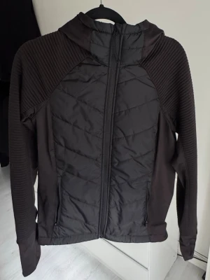 Svart Träningsjacka med huva - Snygg svart träningsjacka ribbade ärmar samt huva. Jackan har dragkedja framtill och en sportig look. Perfekt för dig som gillar stilrena och funktionella plagg.