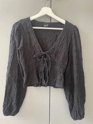 Svart structured top med knytning - Super snygg svart blus från Gina Tricot i crinkle-material med V-ringning och knytning framtill. Blusen har långa puffiga ärmar och en croppad passform💕Toppen är endast använd ett få tal gånger och säljs pga för liten