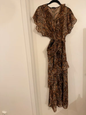 Zara Brun långklänning med ormmönster - Säljer en flowy långklänning i brunt med ormmönster och volangdetaljer. Klänningen har v-ringning, korta ärmar och omlottmodell med knyt i midjan. Perfekt för dig som vill sticka ut med en trendig och djurinspirerad look.