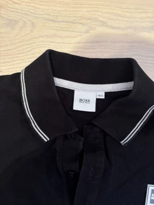 Svart BOSS pikétröja med vit kant (14 år) - Svart t-shirt från BOSS med klassisk pikékrage och vita ränder längs krage och ärmslut. T-shirten har knappslå framtill och en diskret BOSS-logga på bröstet. Tillverkad i mjuk bomull för en clean och stilren look.