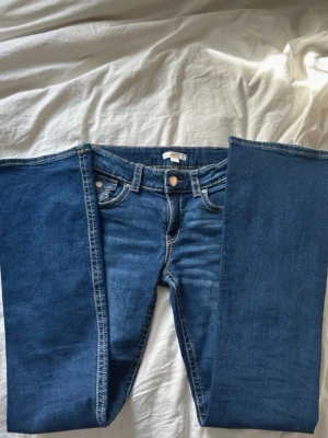 Blå bootcut jeans med fickdetaljer - Säljer ett par klassiska blå bootcut jeans med kontrastsömmar och snygga fickor med lock och knapp bak. Jeansen har fem fickor, bälteshällor och stängs med knapp och dragkedja. Perfekta till en avslappnad och trendig look. Storlek 170!
