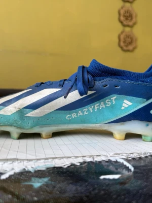 Blå Adidas Crazyfast fotbollsskor - Säljer ett par blå Adidas Crazyfast fotbollsskor med vita detaljer och klassiska tre ränder på sidan. Skorna har snörning, låg profil och en strumpliknande krage för extra stöd. Yttermaterialet är syntetiskt och sulan har fasta dobbar för spel på gräs.