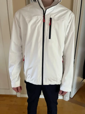 Vit vindjacka från Helly Hansen - Vind- och vattentät jacka från Helly Hansen med Helly Tech Protection-teknologi. Jackan har en stilren design med dragkedja framtill och flera fickor med dragkedja. Kragen är hög och har en mjuk insida för extra komfort. Ärmsluten är justerbara med kardborreband och nederkanten kan dras åt med dragsko för en anpassad passform. Logotypen är synlig på kragen och ärmen.  Modellen på bilden är 180 väger 70kg Den perfekta vårjackan Storlek M Har bara testat den  Hör av er vid minsta fundering!