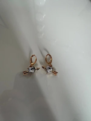 Guldörhängen med hjärtformad sten - Snygga örhängen i guldfärg med hjärtformad klar sten och pil-detalj. Hängande modell som ger en lekfull och trendig look. Perfekta för dig som gillar unika accessoarer med lite extra bling.