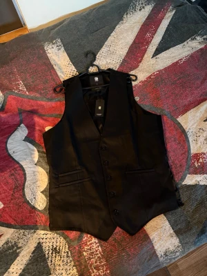 Svart kostymväst från G-Star RAW - Snygg svart kostymväst från G-Star RAW med klassisk V-ringning och fem knappar framtill. Västen har två fickor och är tillverkad i ett stilrent, lätt glansigt material som ger en dressad look. Perfekt att bära över skjorta för en clean och modern stil.