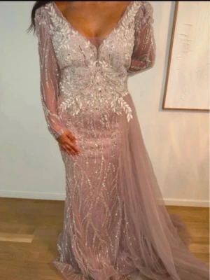 Lång glittrig klänning med spets - Elegant långklänning i ljusrosa med glittriga detaljer och spetsmönster. Klänningen har långärmade meshärmar, v-ringning och ett skirt släp. Perfekt för bal eller fest. 