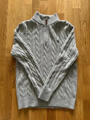 Grå kabelstickad tröja Polo Ralph Lauren - Säljer en grå kabelstickad tröja från Polo Ralph Lauren med halv dragkedja och hög krage. Tröjan har ribbade muddar och den klassiska broderade loggan på bröstet. Perfekt för dig som gillar stilren och tidlös design.