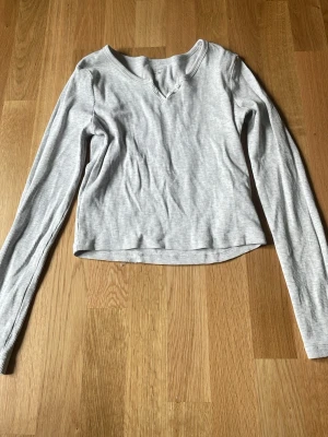 Ljusgrå ribbad långärmad topp - Säljer en ljusgrå ribbad långärmad topp med v-ringning. Toppen har en enkel och clean design, perfekt att matcha med olika stilar. Tillverkad i mjuk bomullsmix för skön känsla hela dagen.