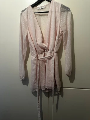 Ljusrosa omlottblus från NA-KD - Söt ljusrosa omlottblus från NA-KD med diskret prickigt mönster. Blusen har långa ärmar, knytband i midjan och ett tunt, lätt material som ger en luftig känsla. Perfekt för att skapa en romantisk och trendig look.