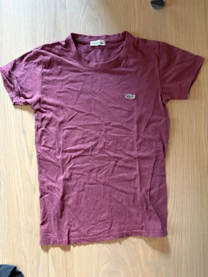 Vinröd t-shirt från Lacoste - Snygg vinröd t-shirt från Lacoste med klassisk krokodillogga broderad på bröstet. T-shirten har rund halsringning och korta ärmar. Tillverkad i mjuk bomull som känns skön mot huden. Perfekt för en avslappnad och stilren look.