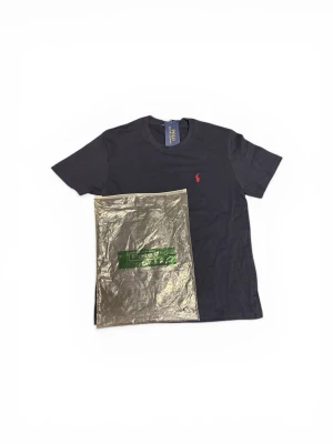 Polo Ralph Lauren T-shirt 👕 - Produkt: Polo Ralph Lauren T-shirt 👕 *   Färg: Mörkblå 🌑 *   Material: 100% bomull 🧶 *   Design: Klassisk crew neck-design med den ikoniska Polo-hästlogotypen i rött broderat på bröstet 🐎. *   Detaljer: Originaletiketten är kvar, vilket tyder på att den är ny. Förpackningen visar att den är förseglad. *   Storlek: M (passar som S) 📏 *   Skick: Helt ny 💯 *   Perfekt för: Vardagskläder, sport eller en avslappnad look. En tidlös klassiker! ✨  Hashtags: #PoloRalphLauren #Tshirt #Fashion #NewWithTags