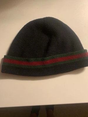 Svart mössa från Gucci med ränder - Säljer en svart ribbstickad mössa från Gucci med klassiska gröna och röda ränder runt kanten. Mössan är i mjukt material och har en enkel, stilren design som passar perfekt till höst och vinter.