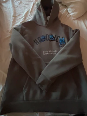 Hoodrich Hoddie - Säljer en brun hoodie från HOODRICH med stor huva och magficka. På bröstet står det HOODRICH i blå och svart text samt ett mindre tryck i vitt under. Hoodien har en avslappnad passform och är gjord i mjukt material, perfekt för chill dagar.