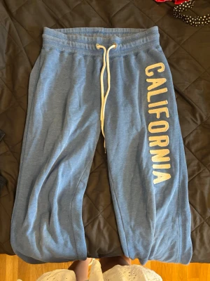 Blå mjukisbyxor med CALIFORNIA-tryck - Säljer ett par blå mjukisbyxor med vitt snöre i midjan och stort vitt CALIFORNIA-tryck längs ena benet. Byxorna har resår i midjan och vid bensluten för extra komfort. Perfekta för chill dagar eller när du vill ha en avslappnad stil.