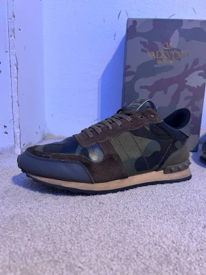 Valentino rockrunners - Säljer ett par snygga sneakers från Valentino med camomönster i grönt och svart. Skorna har detaljer i mocka och läder, samt nitar på hälen för en cool touch. Låg modell med klassisk snörning och bekväm sula. Perfekta för dig som vill sticka ut.