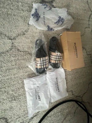 Burberry sandaler med rutigt band - Säljer ett par Burberry sandaler med klassiskt beige, svart och rött rutigt band över foten. Sulan är svart och sandalerna har en stilren och modern design. Perfekta för sommaren och ger en lyxig känsla till din outfit. Kommer med originalförpackning och dustbags.
