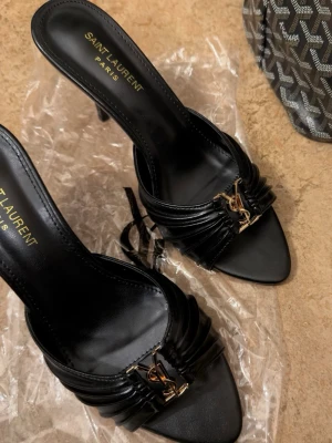  Svarta klackar i lyxig design 🖤 - Säljer ett par eleganta svarta klackar i läder med öppen tå och hög klack. Skorna har fina veckade detaljer framtill och en stilren guldfärgad detalj som ger en exklusiv look.  Endast använda en gång i mycket fint skick. Perfekta till fest, middagar eller när du vill ha en uppklädd, lyxig outfit.  • Stilrena & trendiga • Ger en designer-inspirerad look • Bekväma om du är van vid klackar