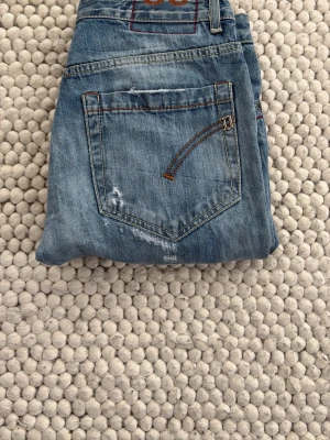 Dondup jeans - Säljer ett par blå jeans med smal passform och slitningar på både fram- och baksidan. Jeansen har klassiska fem fickor och coola detaljer med orange sömmar. Materialet är denim och färgen är ljusblå med distressed look. INGA SKADOR ELLER SÖNDRIGA IGENOM! 