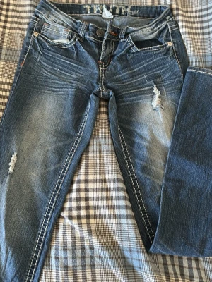 Blå slitna bootcut jeans - Säljer ett par blå bootcut jeans med slitningar och ljusa tvättade partier. Jeansen har vita kontrastsömmar och klassisk femficksmodell. Materialet är denim i bomull och passformen är något utsvängd nedtill. En fin och detaljerad bakficka. Jeansen några få orangea sömmar. 