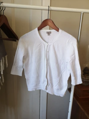 Fin kofta  - Klassisk vit kofta från med trekvartsärmar. Koftan har knäppning framtill med små vita knappar och ribbade muddar vid ärmslut och nederkant. Perfekt att slänga över en topp för en clean look. Reserv knappen finns fortfarande kvar på lappen. St M men passar st S