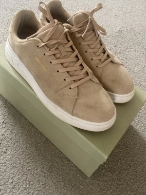 Beige sneakers från Iguasu - Stilrena beige sneakers från Iguasu med ovandel i mockaliknande material och klassisk vit sula. Skorna har lågt skaft, matchande beige snörning och diskret logga i guld på sidan. Perfekta för en clean och avslappnad look.