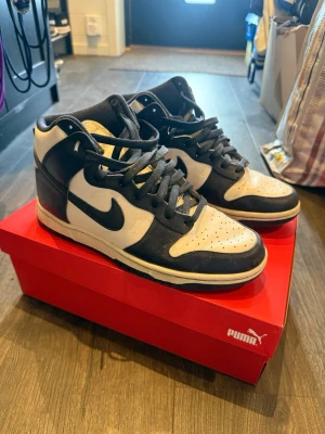 Nike Dunk High Retro svart/vit - Nike Dunk High Retro sneakers i svart och vitt läder med klassisk Swoosh-logga på sidorna. Högt skaft och perforerad tå för extra ventilation. Stilren och tidlös modell som passar perfekt till streetwear och casual outfits.