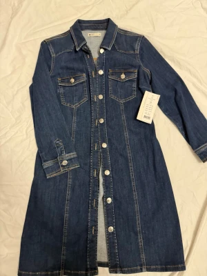 Mörkblå jeansklänning från Gina Tricot - Snygg mörkblå jeansklänning som är knälång från Gina Tricot med långa ärmar, knappar hela vägen framtill och två bröstfickor med lock. Klänningen har klassisk skjortkrage och kontrastsömmar som ger en cool denimlook. Liten i storlek strechmaterial 