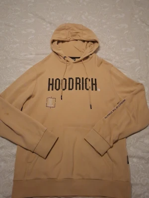 Beige hoodie från Hoodrich med tryck - Snygg beige hoodie från Hoodrich med stor logga på bröstet och grafiskt tryck på ryggen. Hoodien har huva, ficka och textdetaljer på ena ärmen. Tillverkad i mjukt bomullsmaterial för skön känsla och avslappnad passform.