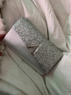 Silverglittrig kuvertväska med kedja - Snygg kuvertväska i silver från LYDC med glittrigt lock och metallkedja som axelrem. Väskan har ett stilrent V-format metallspänne framtill och är tillverkad i ett skimrande material som verkligen sticker ut. Perfekt accessoar för att lyfta din outfit.