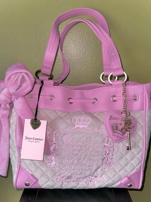 Juicy Couture daydreamer tote rosa väska - Superfin quiltad väska från Juicy Couture i ljusrosa och grått med stora handtag, dekorativ rosett och hjärtformad nyckelberlock. Väskan har broderad logga och detaljer i silver. Perfekt för dig som gillar en söt och trendig look.