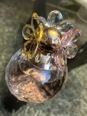 Parfymflaska med blommig kristallkork - Säljer en superfin parfymflaska i ljusrosa glas med slipad yta. Korken är dekorerad med stora, genomskinliga blommor i plast med inslag av gult och rosa. Flaskan har en lyxig och lekfull design som verkligen sticker ut i badrumshyllan.