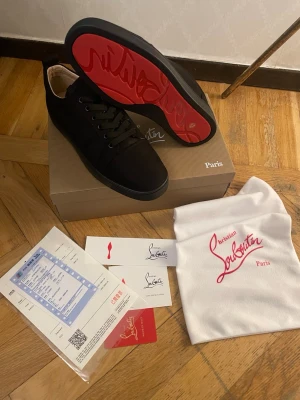 Svarta Christian Louboutin sneakers - Snygga svarta sneakers från Christian Louboutin med klassisk röd sula och diskret logga. Skorna har lågt skaft, svarta skosnören och är tillverkade i mocka. Perfekta för dig som vill ha en exklusiv och stilren look.