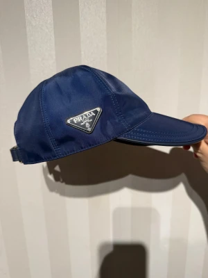 Mörkblå keps från Prada - Snygg mörkblå keps från Prada med klassisk böjd skärm och justerbar rem baktill. Kepsen är tillverkad i nylon och har Pradas ikoniska metalltriangel på sidan. Perfekt för dig som vill ha en stilren accessoar med lyxig känsla.