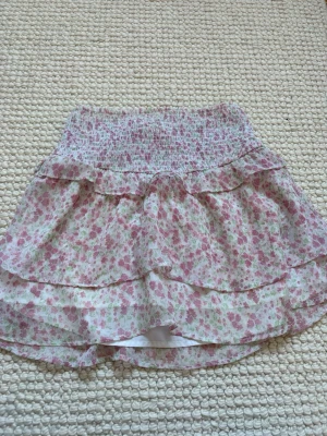 Blommig kortkjol med volang - Söt kortkjol med blommigt mönster i rosa och grönt på vit bakgrund. Kjolen har bred smock i midjan och volangdetaljer som ger ett luftigt och romantiskt uttryck. Perfekt för dig som gillar en feminin och lätt stil. Stretchig så passar även om du är större än en S. 