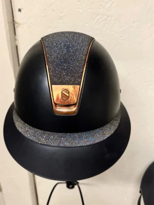 Svart ridhjälm med glitter och koppar - Snygg marinblå ridhjälm från Samshield med glittrande detaljer på toppen och skärmen samt kopparfärgad emblem och kant. Hjälmen har en modern och stilren design, perfekt för dig som vill sticka ut i stallet.