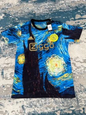 Ajax x Van Gogh matchtröja Adidas - Unik Ajax matchtröja från Adidas med Van Gogh-inspirerat motiv i blått, gult och svart. Tröjan har Ziggo-logga, Ajax-emblem och AEROREADY-material för extra komfort. Perfekt för fotboll eller träning.