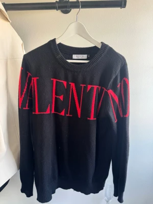 Svart stickad tröja från Valentino - Säljer en svart stickad tröja från Valentino med stort rött VALENTINO-tryck över bröstet. Tröjan har rund halsringning och långa ärmar. Perfekt för dig som vill ha en statement piece i garderoben.