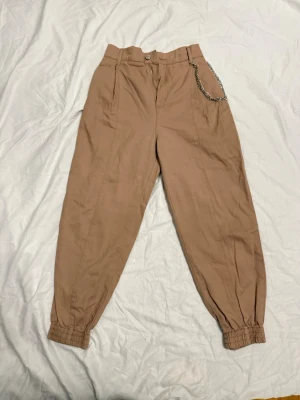Beige cargobyxor med kedja Pull&Bear - Snygga beige cargobyxor från Pull&Bear med avslappnad passform och elastiska muddar vid bensluten. Byxorna har en cool silverfärgad kedja i midjan . Perfekta för en trendig streetwear-look.