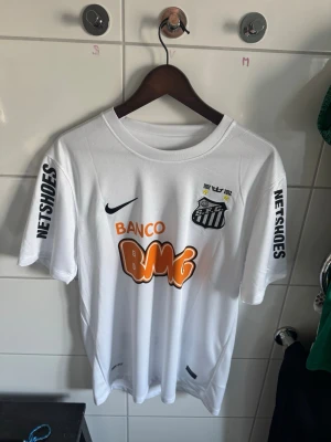 Vit Santos Neymar Jr matchtröja - Säljer en vit Santos FC matchtröja från Nike med Neymar Jr och nummer 11 på ryggen. Tröjan har tryckta sponsorer som BMG och Netshoes samt klubbmärke på bröstet. Korta ärmar och sportig passform, perfekt för fotboll eller träning.
