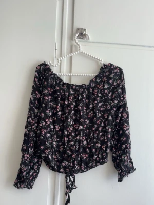 Svart blommig offshoulder-blus - Snygg svart offshoulder-blus med små rosa och vita blommor. Blusen har långa ärmar med resår vid ärmslut och en dragsko nertill för en trendig passform. Perfekt för dig som gillar romantisk och feminin stil.
