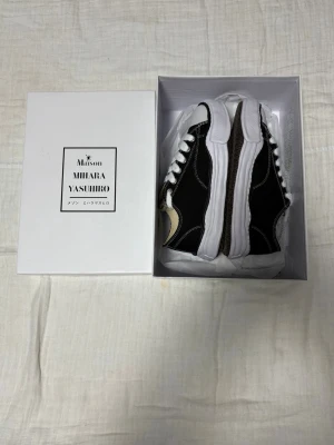 Maison Mihara Yasuhiro svarta sneakers - Svarta sneakers från Maison Mihara Yasuhiro med vit, chunky sula och kontrasterande vita sömmar. Skorna har vit tå, vita skosnören och en unik, vågig design på sulan. Klassisk låg modell med canvasmaterial och cool streetkänsla.
