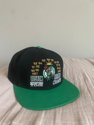 Boston Celtics keps Mitchell & Ness - Snygg svart och grön snapback-keps från Mitchell & Ness med Boston Celtics broderad logga och text. Framsidan har mästerskapsår och Celtics-motiv, baksidan har broderad Mitchell & Ness-logo. Justerbar storlek och platt skärm. Perfekt för basketfans.