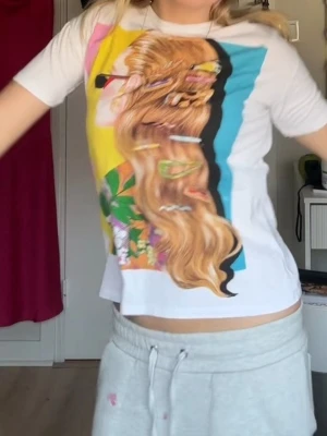 Vit t-shirt med färgglatt tryck Zara - Vit t-shirt från Zara med ett stort färgglatt tryck av en kvinna med långt hår och hårspännen på framsidan. T-shirten har rund hals, korta ärmar och en loose passform. Materialet är mjuk bomull som känns skönt mot huden.