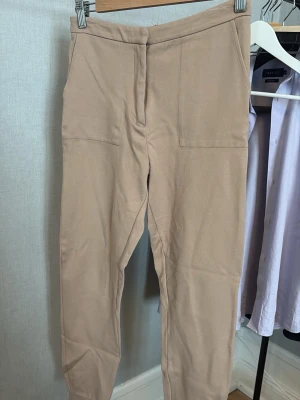 Beige mjukisbyxor med fickor - Snygga beige mjukisbyxor med raka ben och breda muddar vid bensluten. Byxorna har två stora framfickor och en enkel, stilren design. Perfekta för en avslappnad look och tillverkade i mjukt material som känns skönt mot huden.